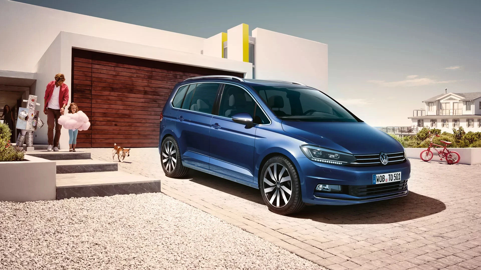 Volkswagen Touran bleu garé devant une maison moderne, avec une famille et un chien devant le garage