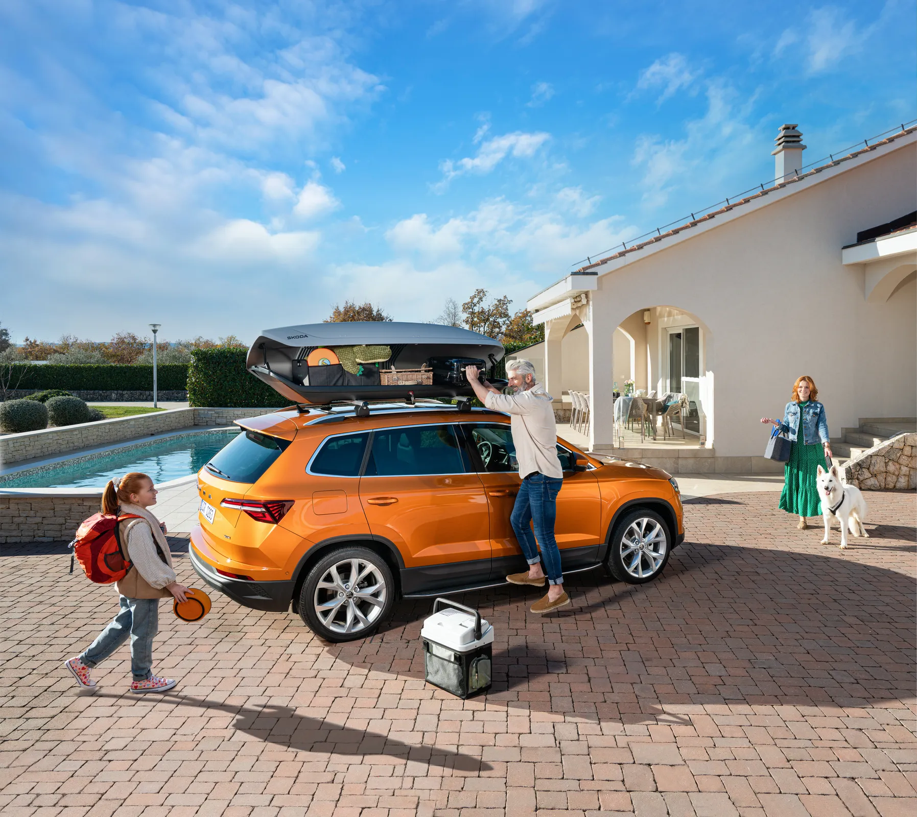 Škoda Karoq orange garé devant une maison, avec coffre de toit et famille qui prépare un départ en week-end