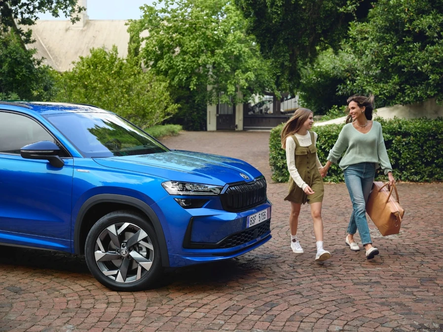 Mère et adolescente souriantes marchant devant un SUV Škoda bleu, prêtes à partir en escapade en famille.